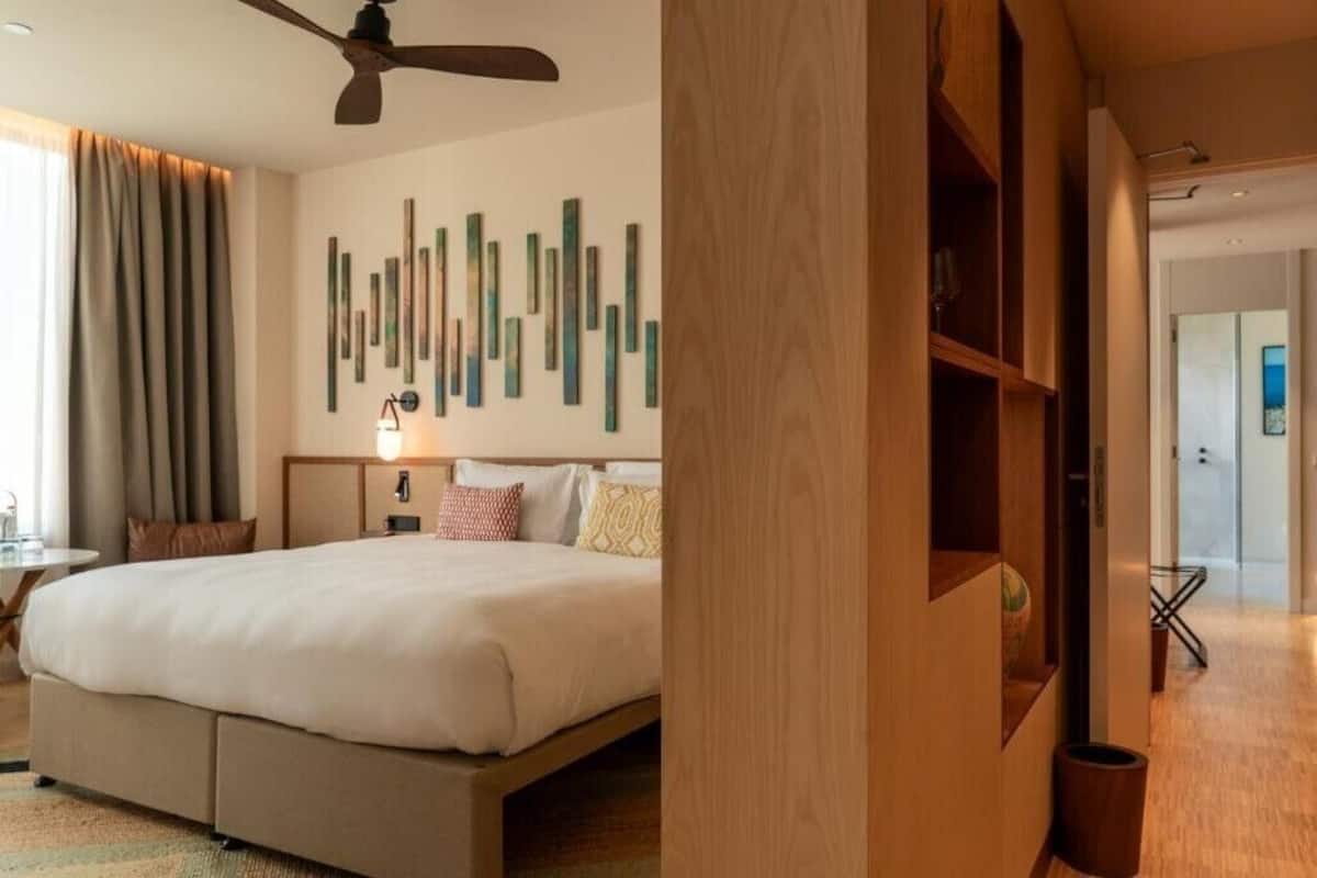 A hotel room with a bed, a ceiling fan, a wall with vertical wooden slats, and a door leading to another room.