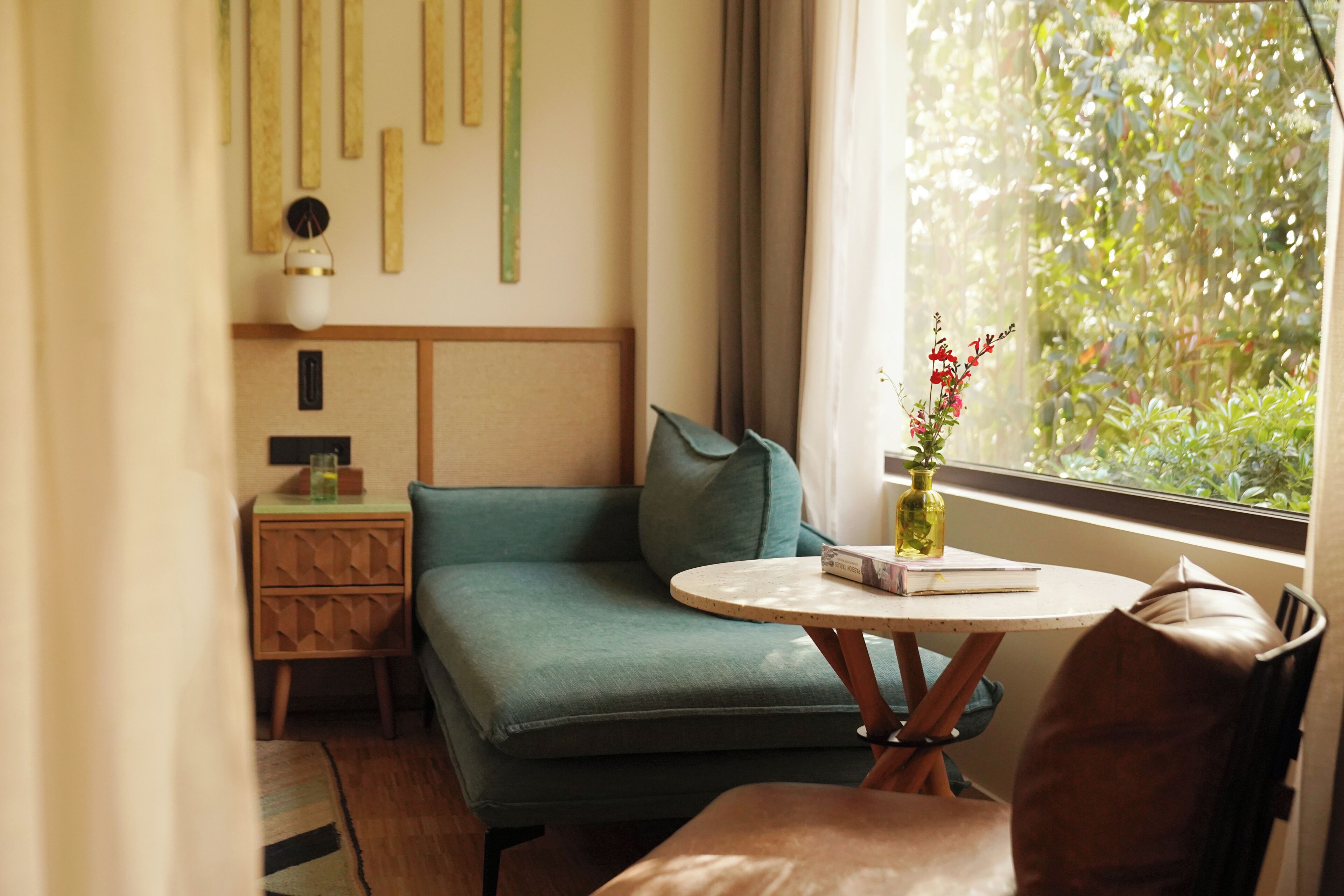 A room with a green sofa, a round table with a vase, a small wooden nightstand, and a window with a view of greenery.