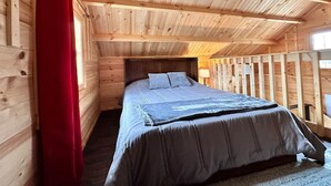 A wooden cabin bedroom with a bed, a red curtain, and a sloped ceiling.