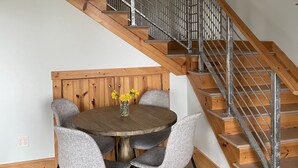 Een trap met houten treden en metalen leuningen, die leidt naar een hoger niveau. Daaronder staat een ronde houten tafel met vier grijze stoelen en een vaas met gele bloemen.