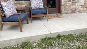 Een stenen huis met een houten veranda, twee houten stoelen met gekleurde kussens, en een hond zichtbaar door een deur.