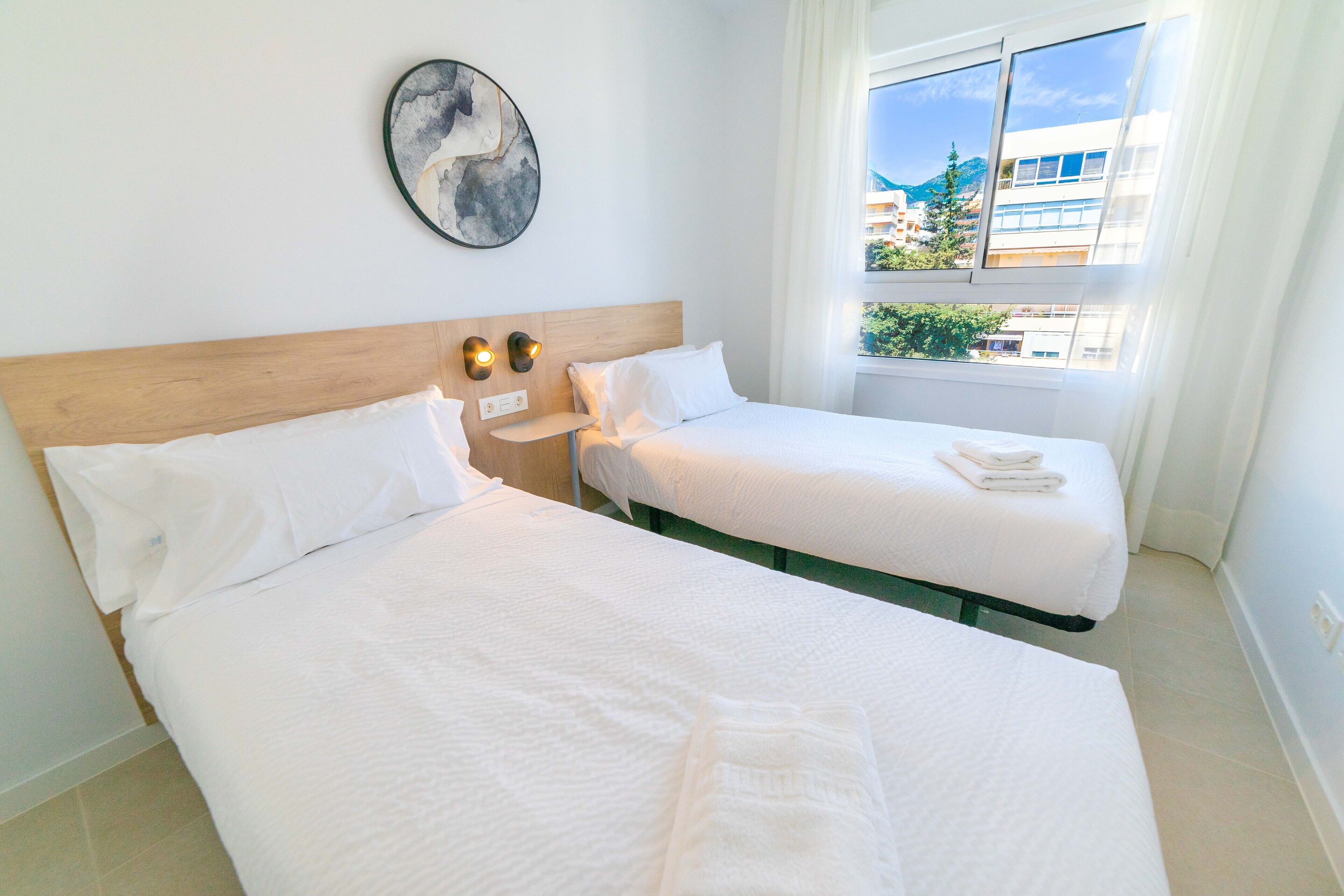 Habitación de hotel con dos camas, cabecera de madera, luces fijadas a la pared, una obra de arte redonda y ventana con vistas a edificios y vegetación.
