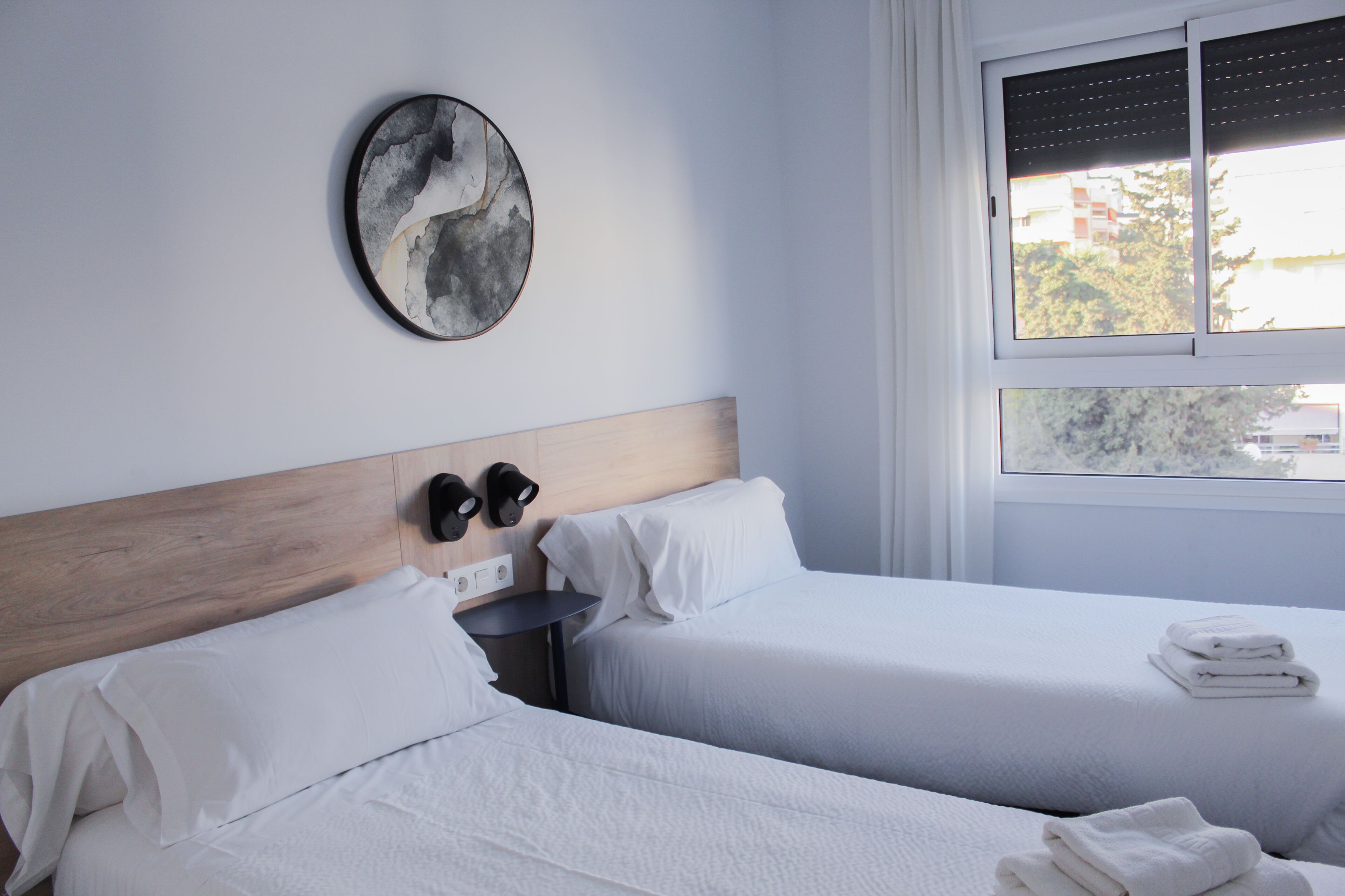 A hotel room with two beds, a wooden headboard, a wall-mounted artwork, a window with blinds, and a view of buildings outside.