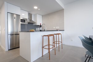 Een moderne keuken met een roestvrijstalen koelkast, een magnetron en een spoelbak. Twee houten krukken en een donkergrijze stoel zijn aanwezig.