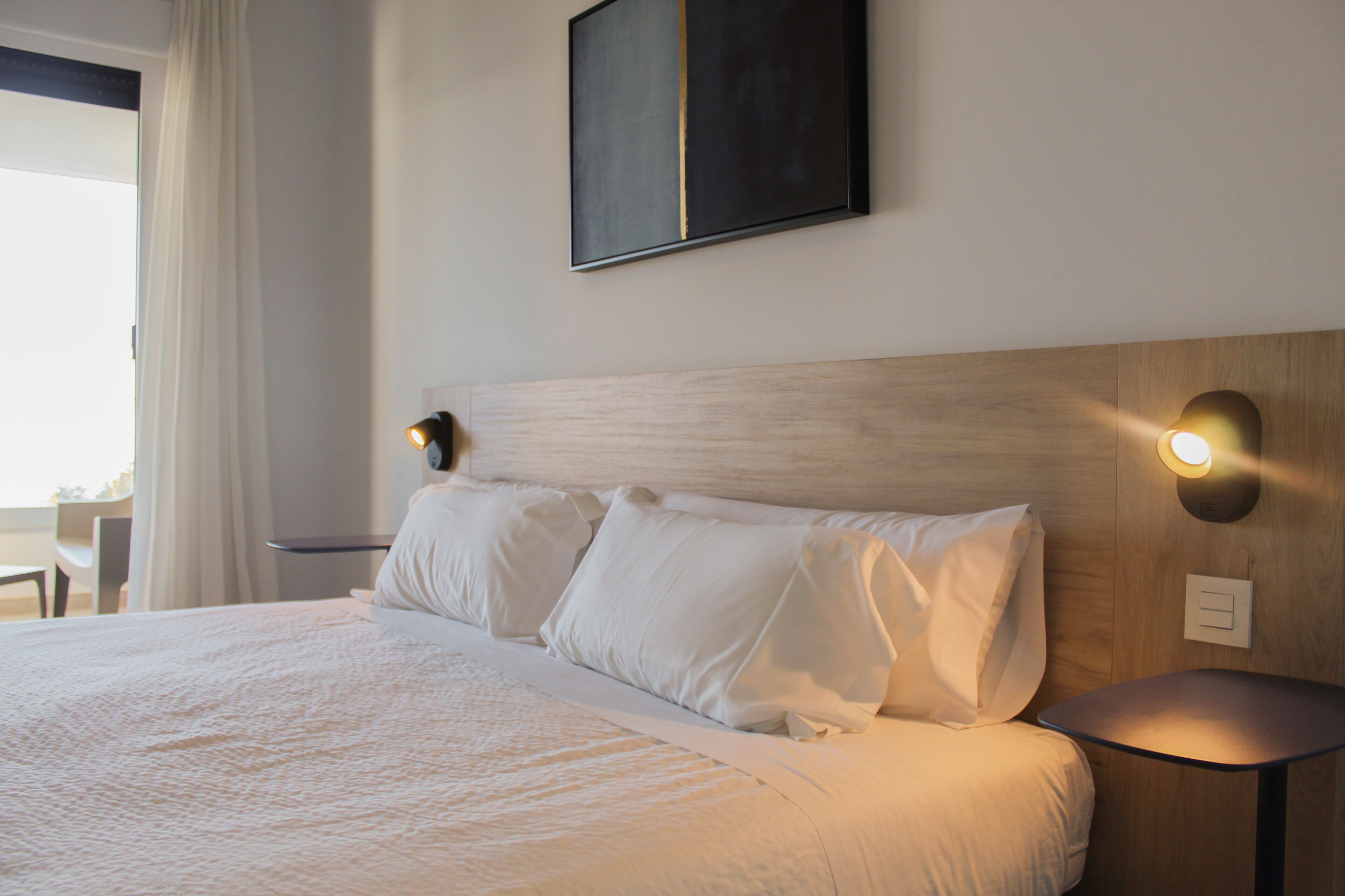 A neatly made bed with white linens, a wooden headboard, and two wall-mounted reading lamps.