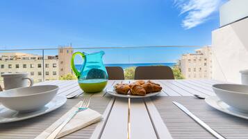 A table set for breakfast with croissants, a pitcher, and cups, overlooking a sea view with buildings in the background.