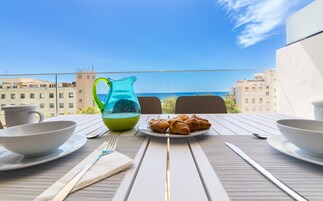 Een ontbijttafel met croissants, een karaf en kopjes, met uitzicht op zee en gebouwen op de achtergrond.
