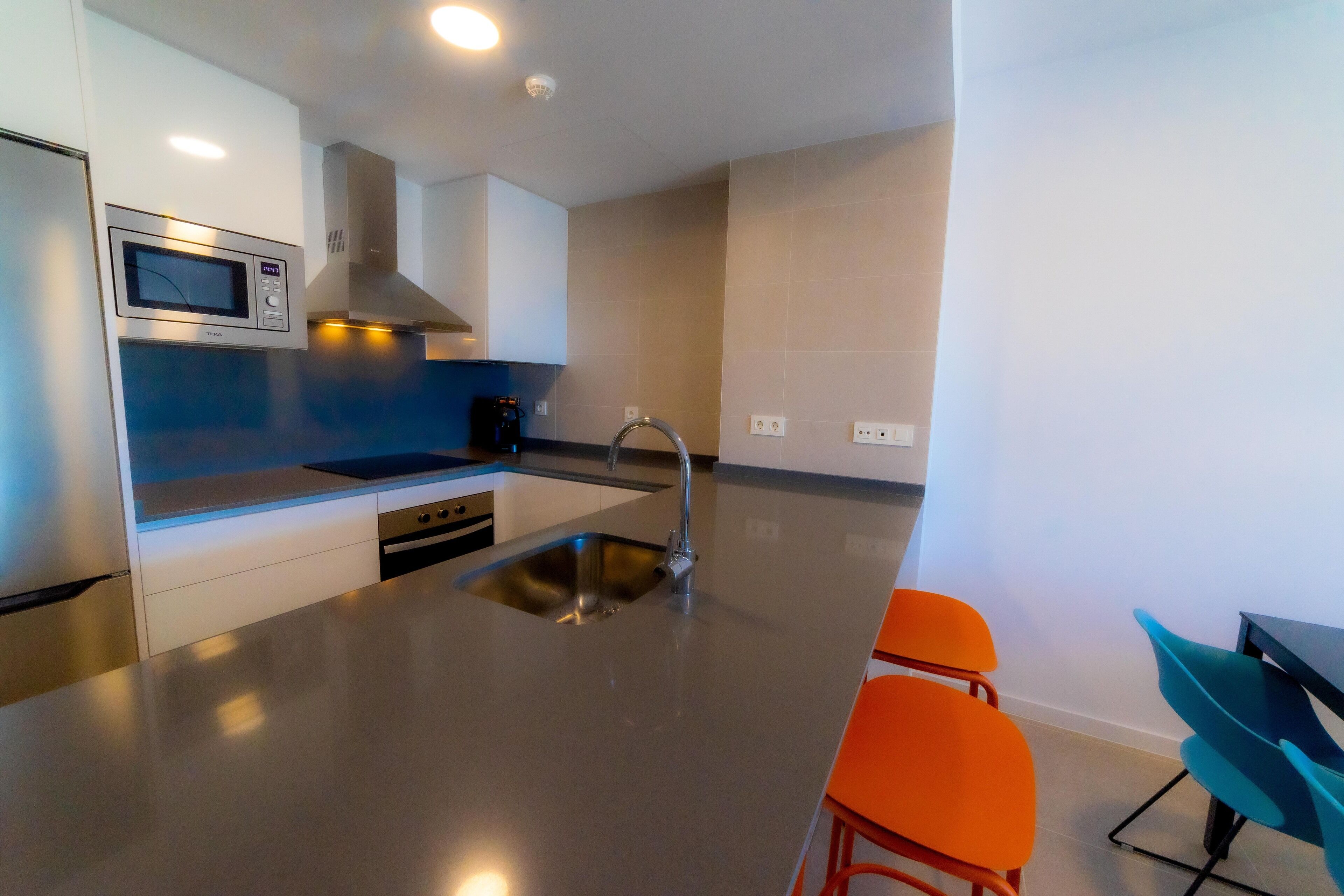 A modern kitchen with a stainless steel sink, a built-in microwave, and a gas stove. There are two orange bar stools and a teal chair.