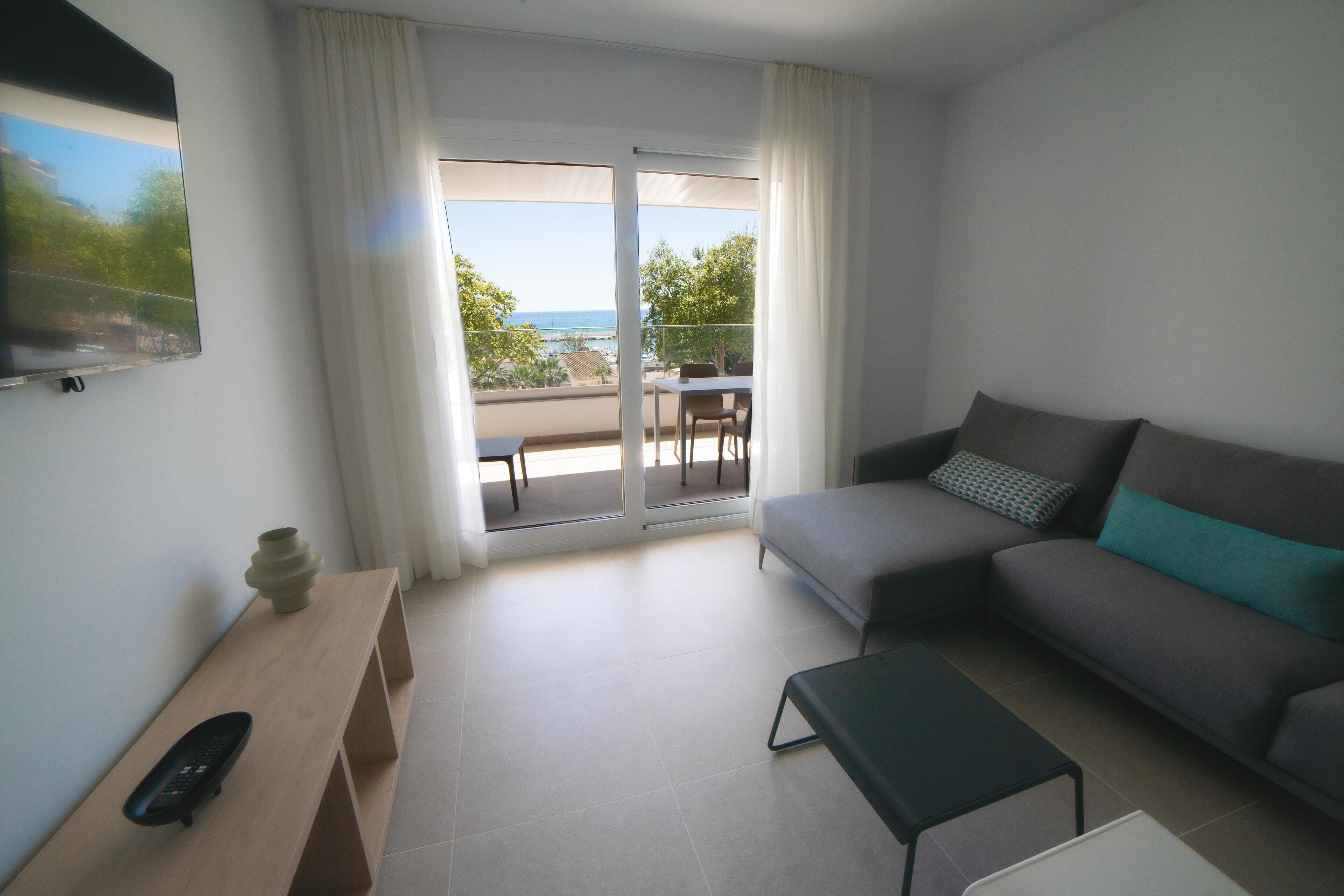 A modern living room with a sofa, a coffee table, a TV, and a balcony with a view of the sea.
