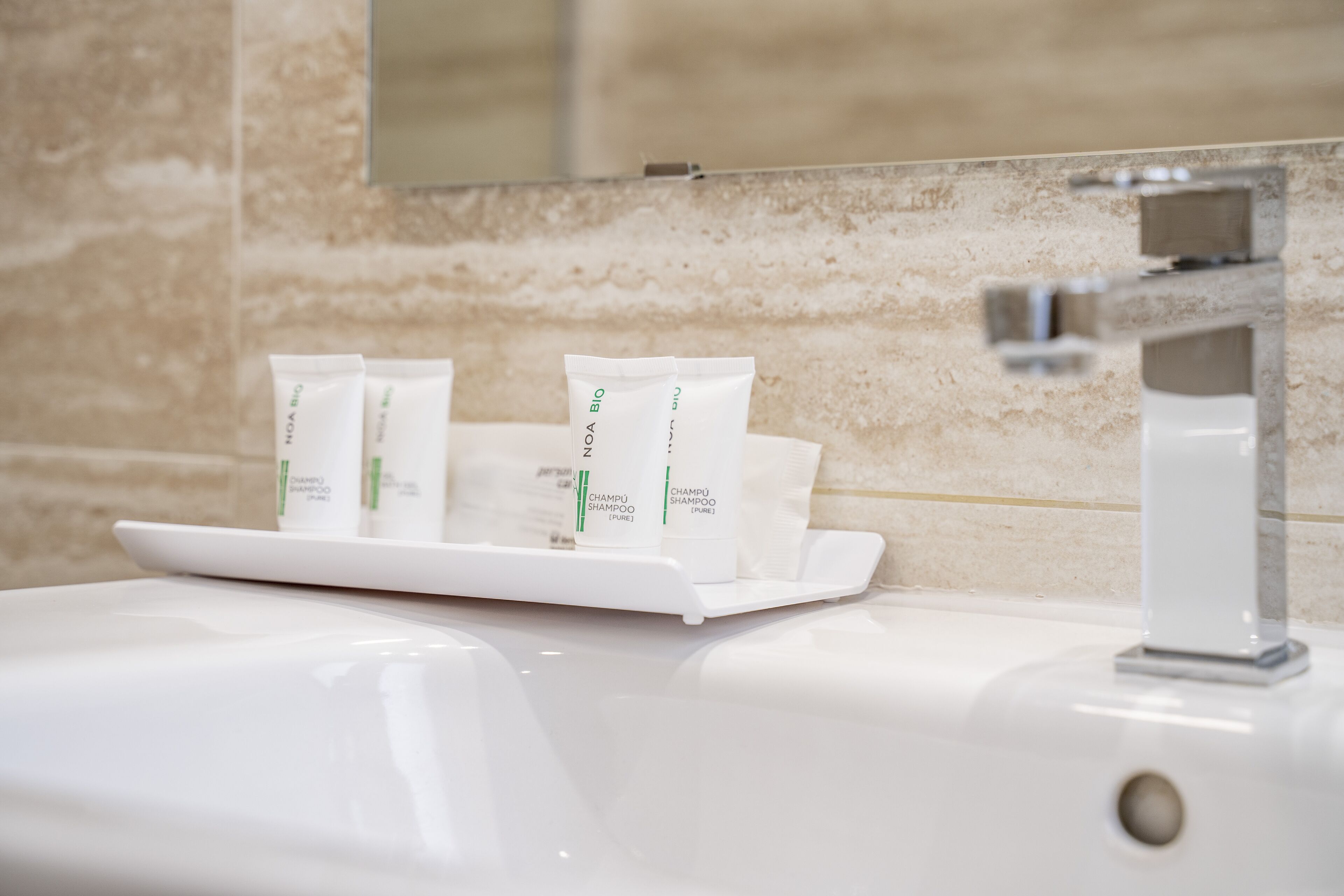 Een wastafel in een badkamer, met daarboven een spiegel en drie flesjes shampoo.