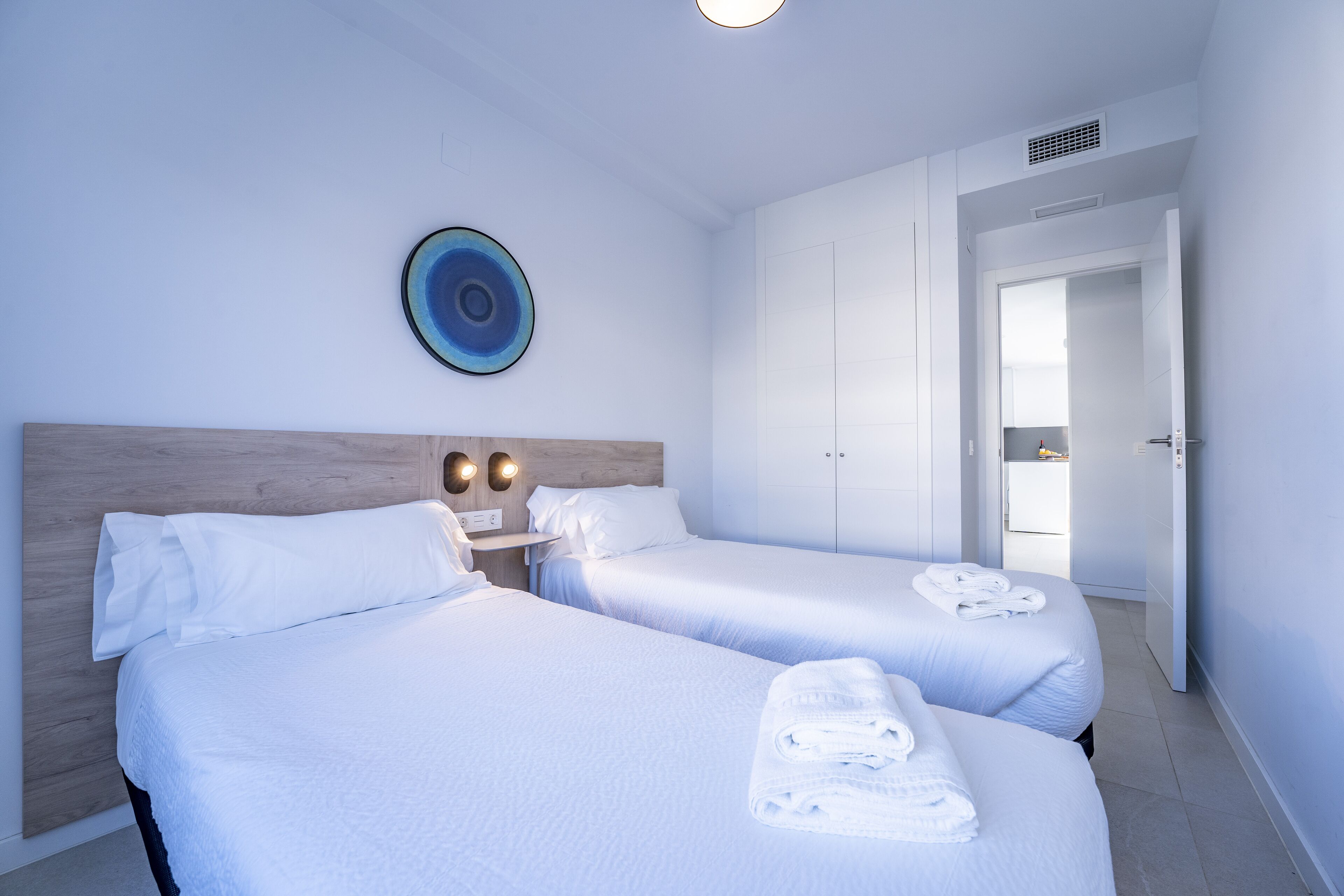 Quarto de hotel com duas camas, cabeceira de madeira, luminária fixada na parede e porta para um banheiro.
