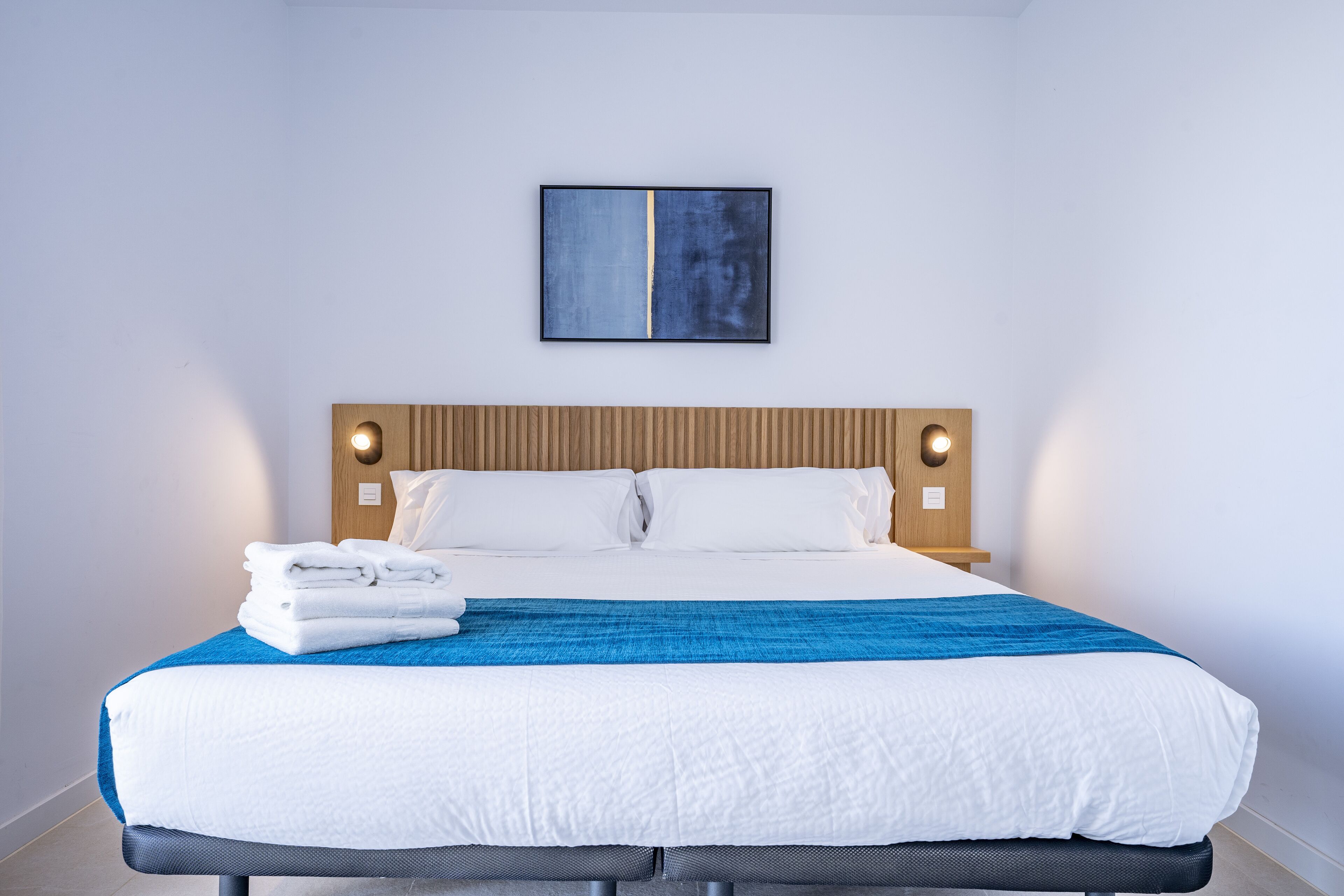 A neatly made bed with white and blue bedding, a wooden headboard, and a single abstract painting on the wall.