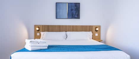 A neatly made bed with white and blue bedding, a wooden headboard, and a single abstract painting on the wall.