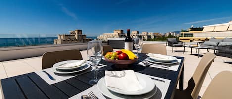 A dining table set with plates, cutlery, and a bowl of fruit, overlooking a coastal cityscape.
