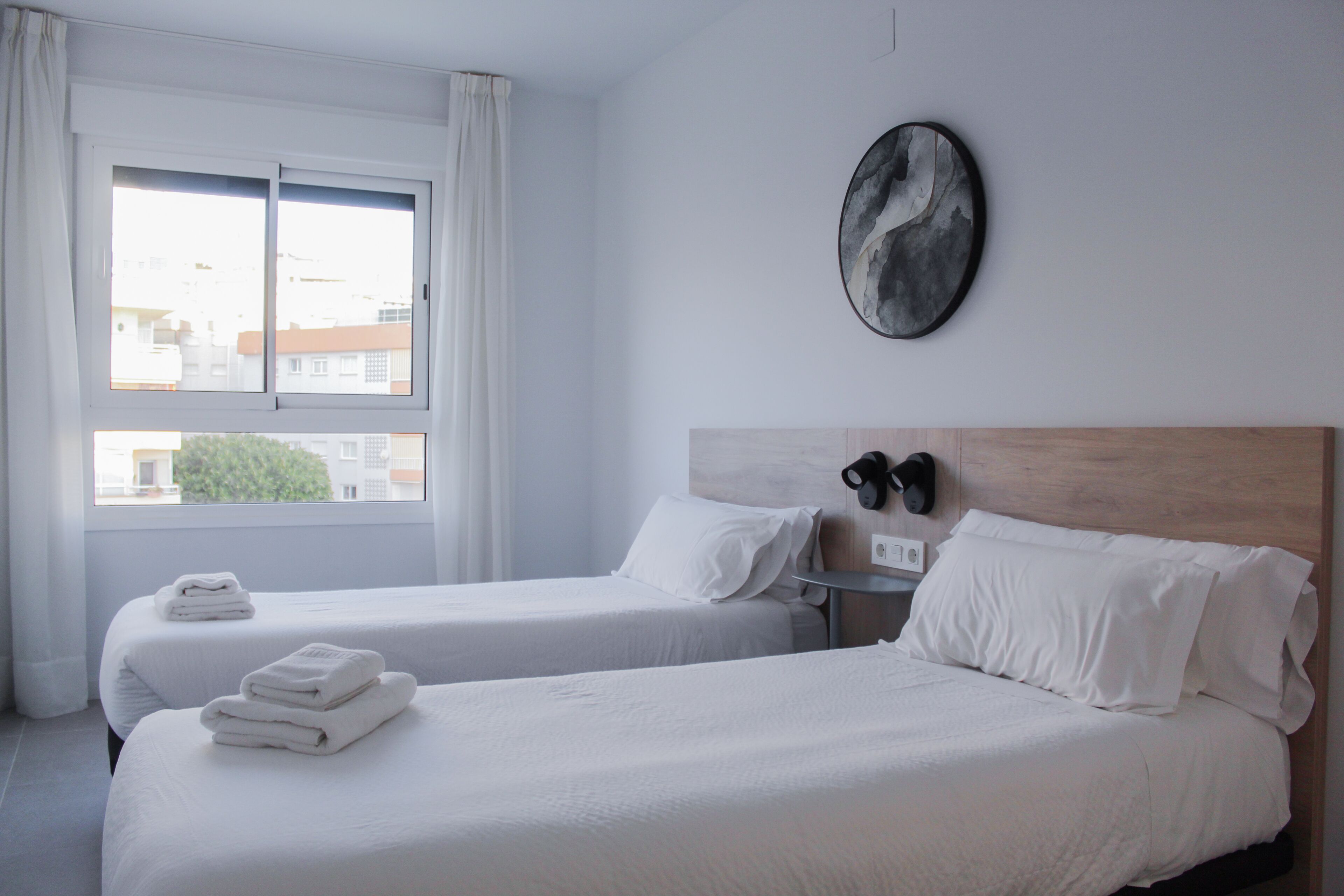 A hotel room with two beds, a large window, a wall-mounted artwork, and a nightstand with a lamp.