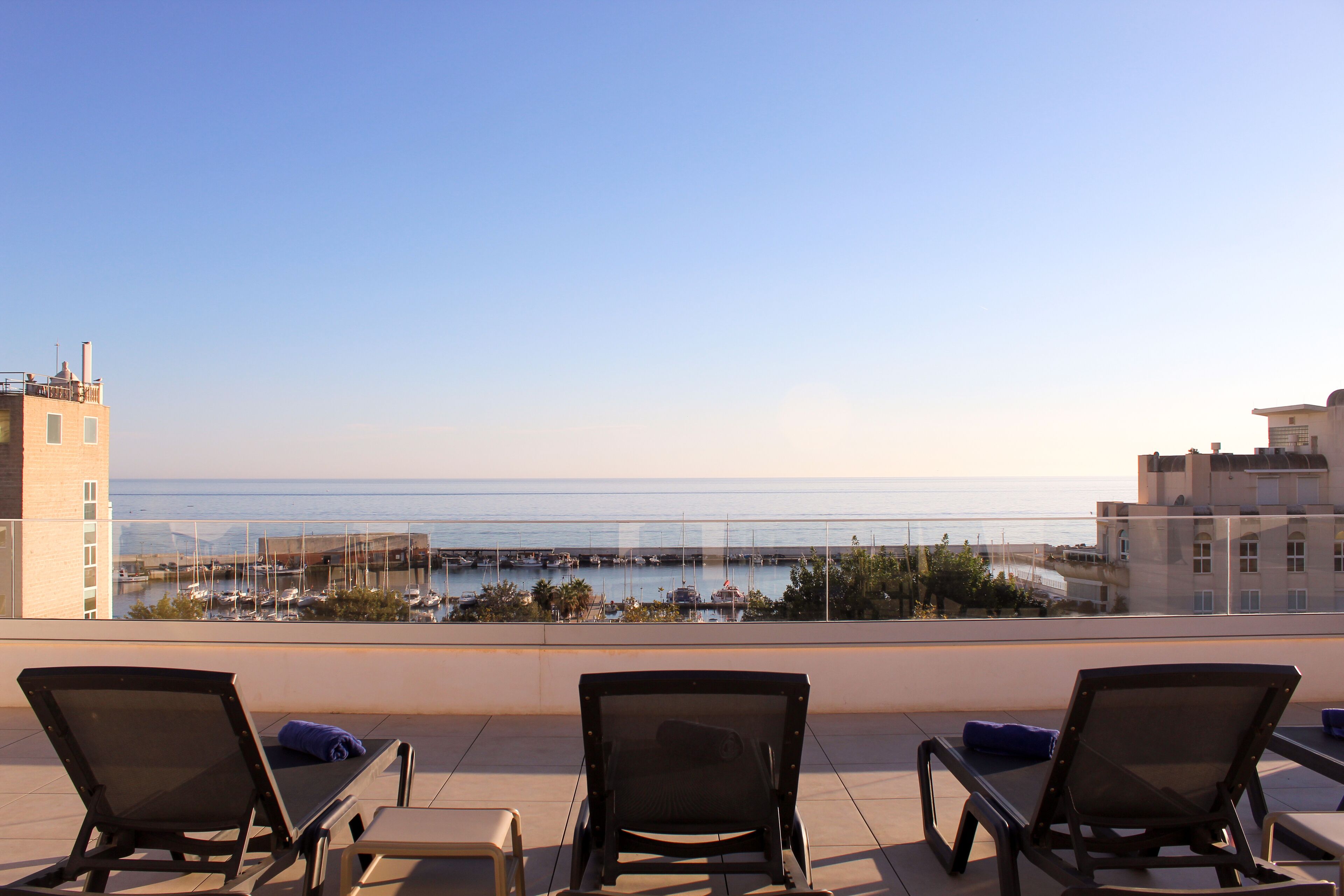 A rooftop terrace with three lounge chairs, a table, and a view of the sea and marina.