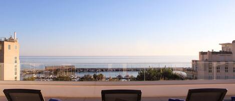 A rooftop terrace with three lounge chairs, a table, and a view of the sea and marina.