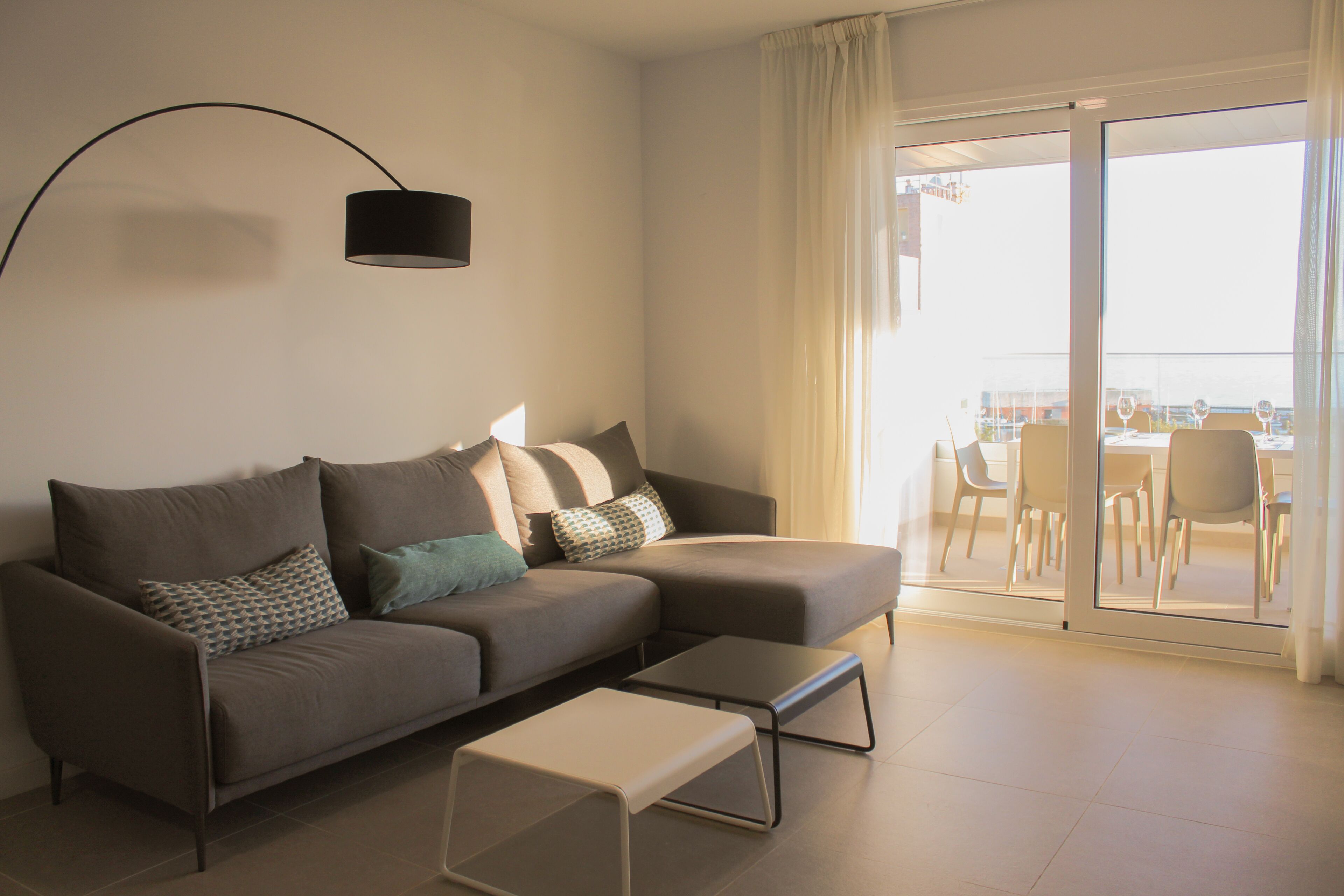 A modern living room with a sofa, a coffee table, and a floor lamp. There is a sliding glass door leading to an outdoor area with chairs and a table.
