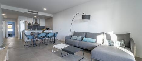 Een moderne woonkamer met een grijze bank, een salontafel en een eethoek met blauwe stoelen.