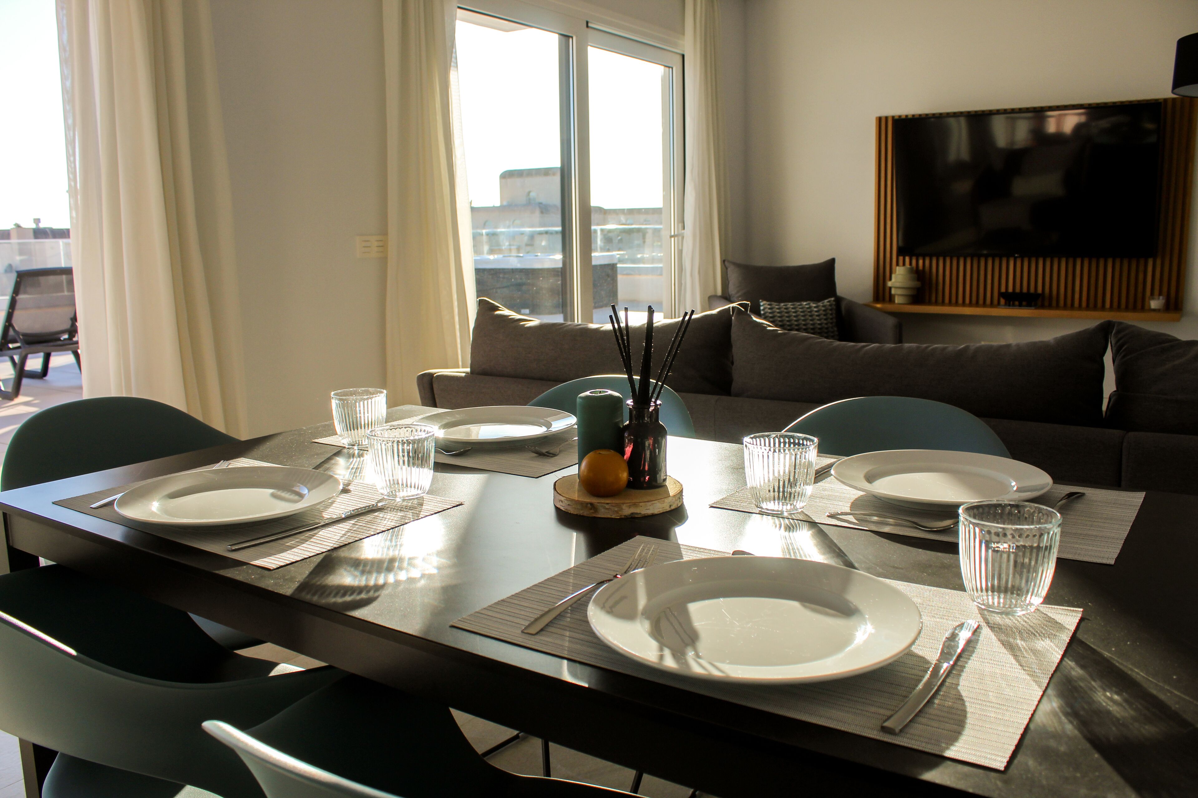 Un comedor con una mesa preparada para cuatro personas, un sofá y un televisor al fondo.