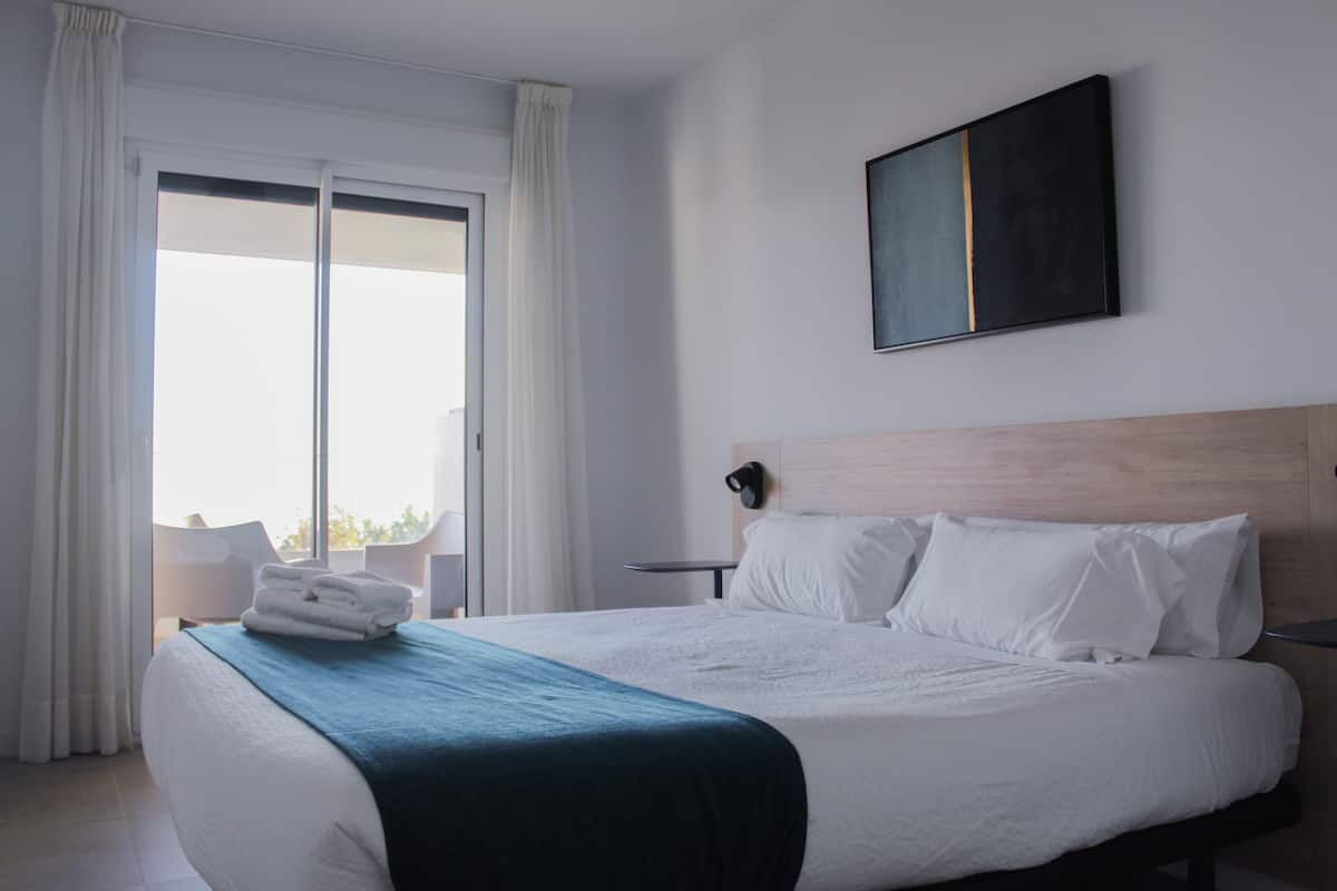 A hotel room with a large bed, a view of the outdoors through a sliding door, a wall-mounted artwork, and a small table with a chair.
