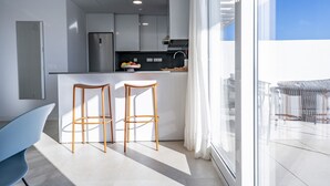 Une cuisine moderne avec des meubles blancs, un réfrigérateur en acier inoxydable et un panier de fruits sur le comptoir. On y trouve deux tabourets en bois près d’une grande fenêtre ornée de rideaux blancs.