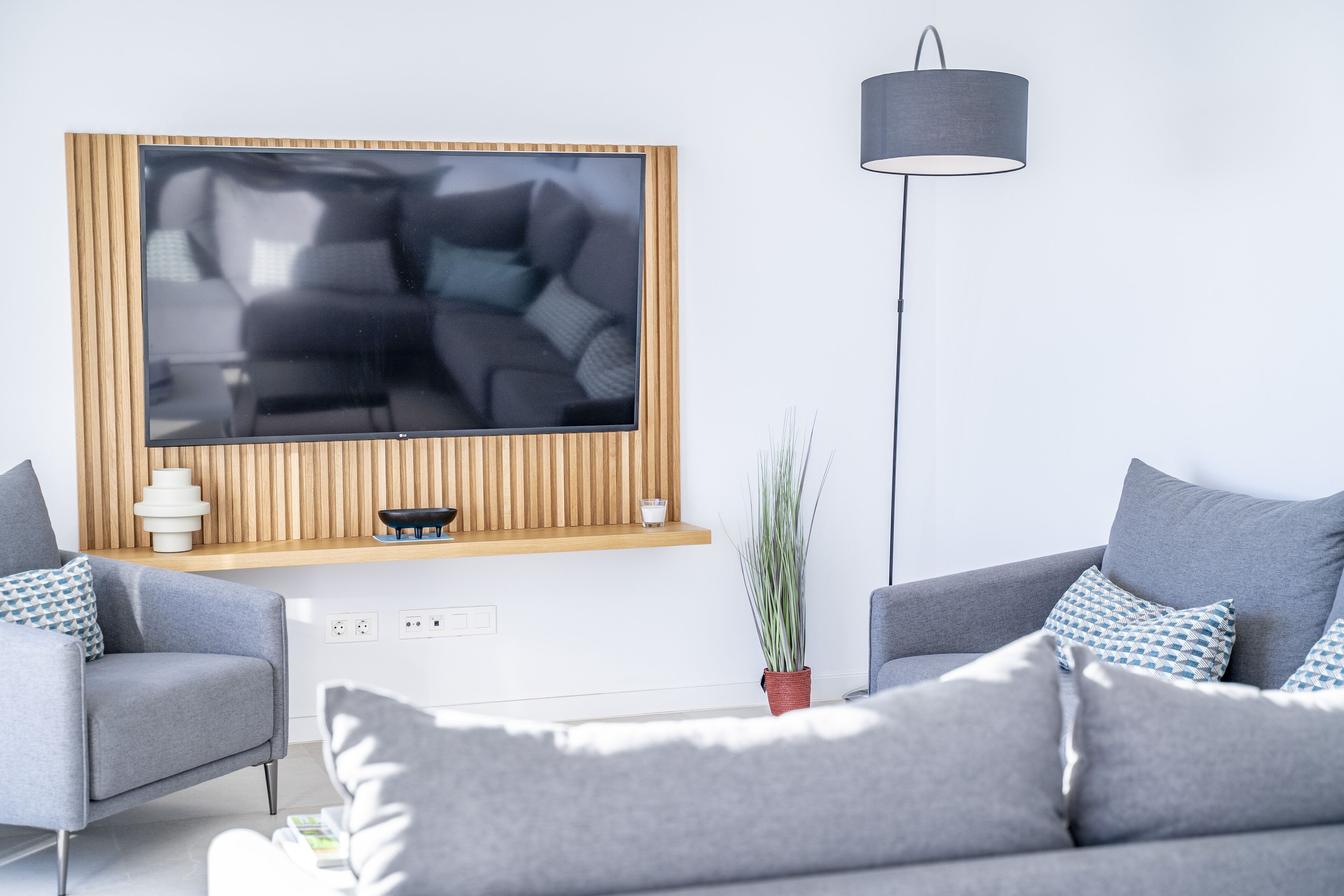 Una sala de estar moderna con un televisor de pantalla plana grande montado en una pared de madera, dos sofás grises con cojines estampados, una lámpara de pie y una planta en maceta.