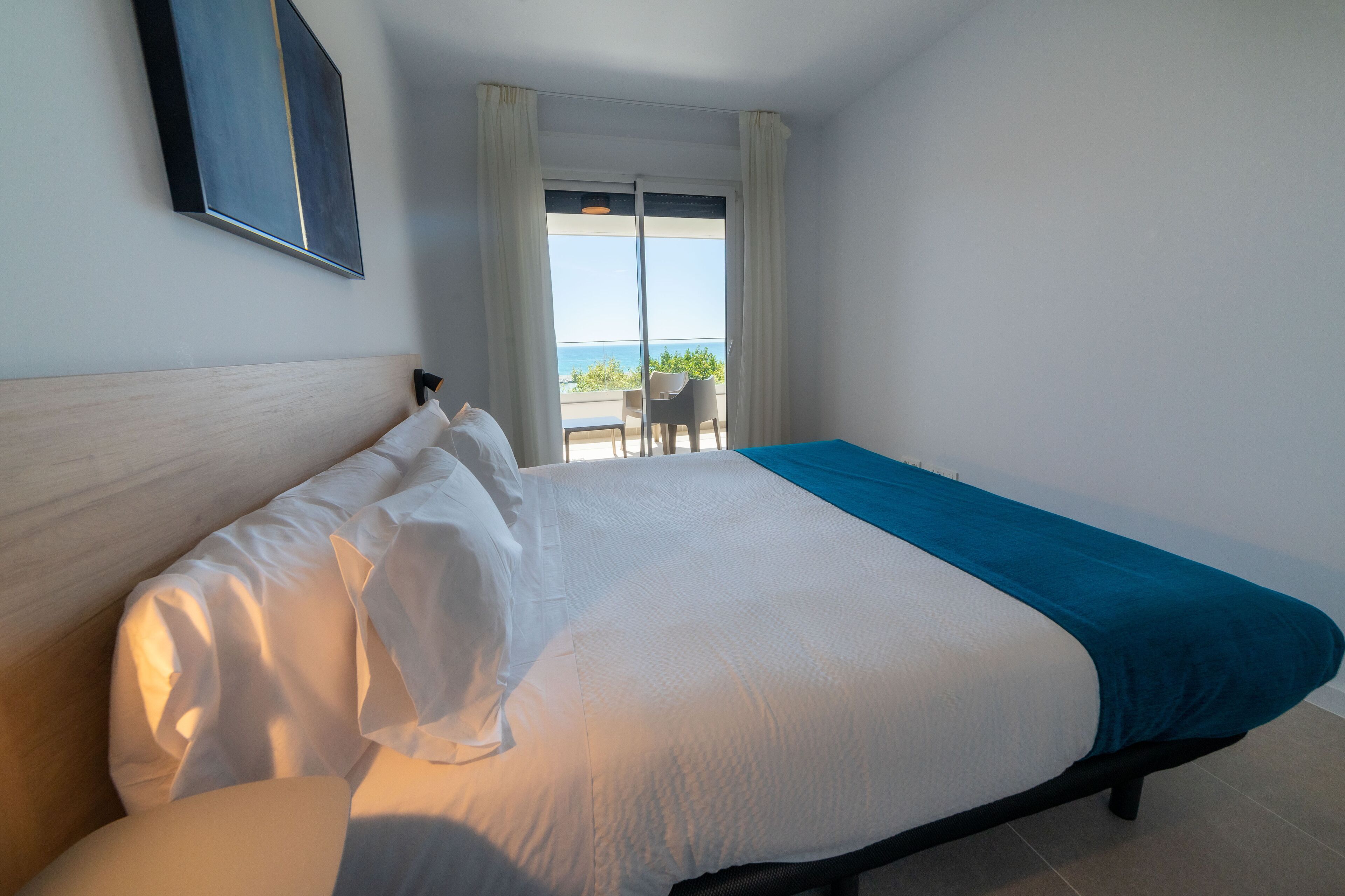 A hotel room with a bed, a view of the sea through the window, and a balcony with a table and chairs.
