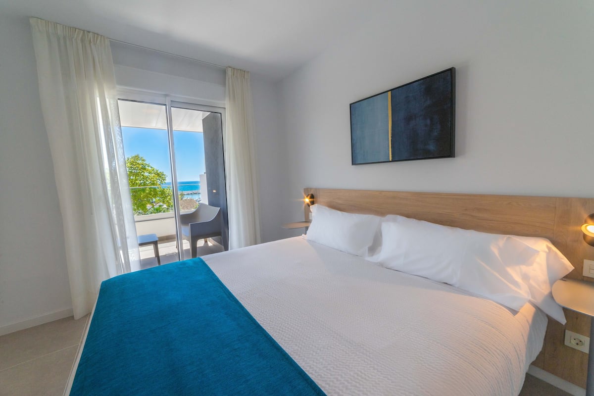 A hotel room with a large bed, a view of the sea through a sliding door, and a painting on the wall.