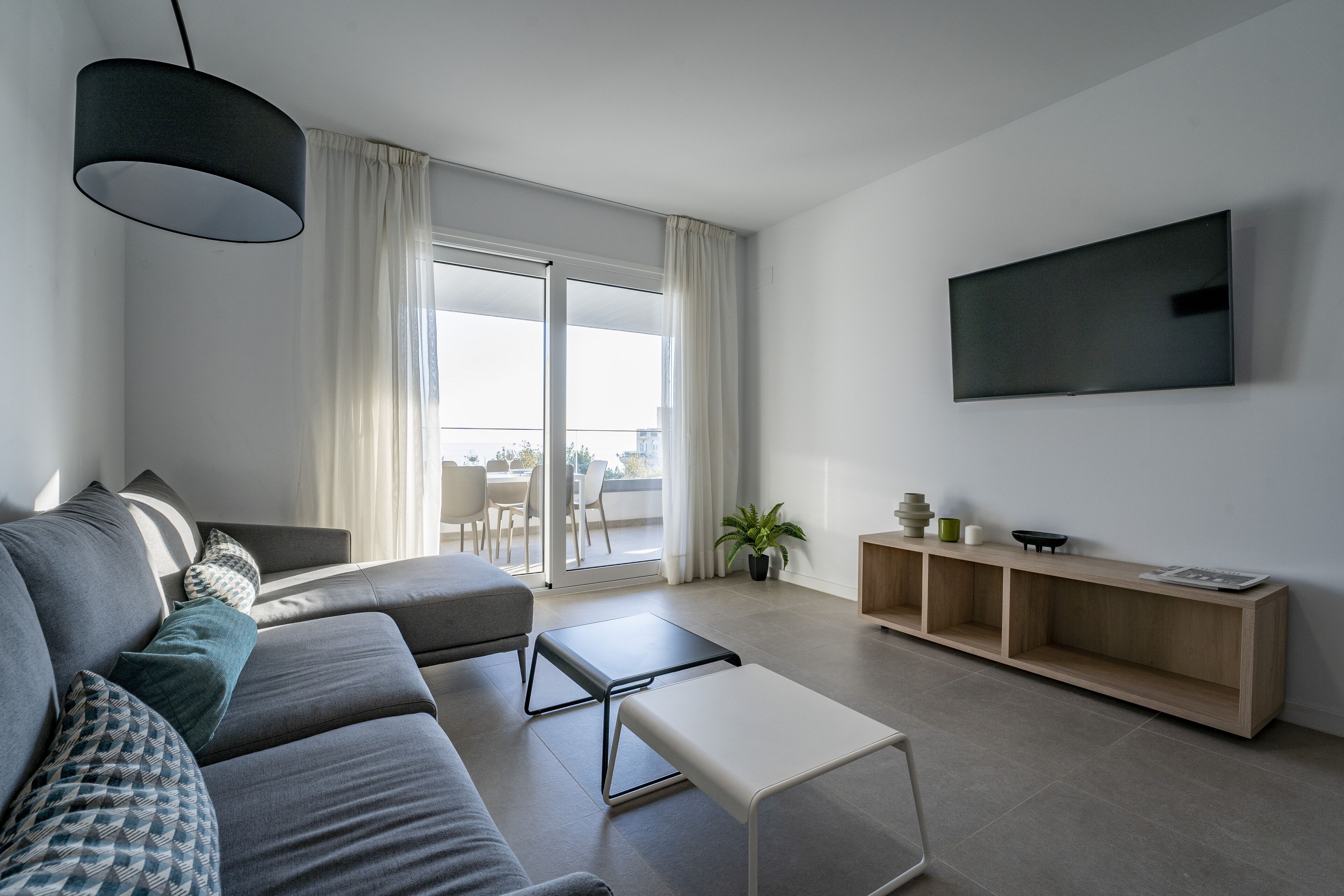 Una sala de estar moderna con un sofá gris, una mesita blanca, un televisor montado en la pared y un ventanal corredero que da a un espacio exterior.
