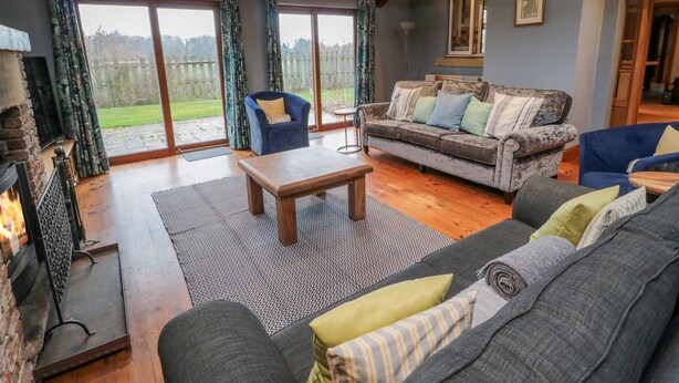 A spacious living room with a fireplace, a sofa set, a coffee table, and a view of the outdoors through large windows. - Ridley Lodge (Morpeth)
