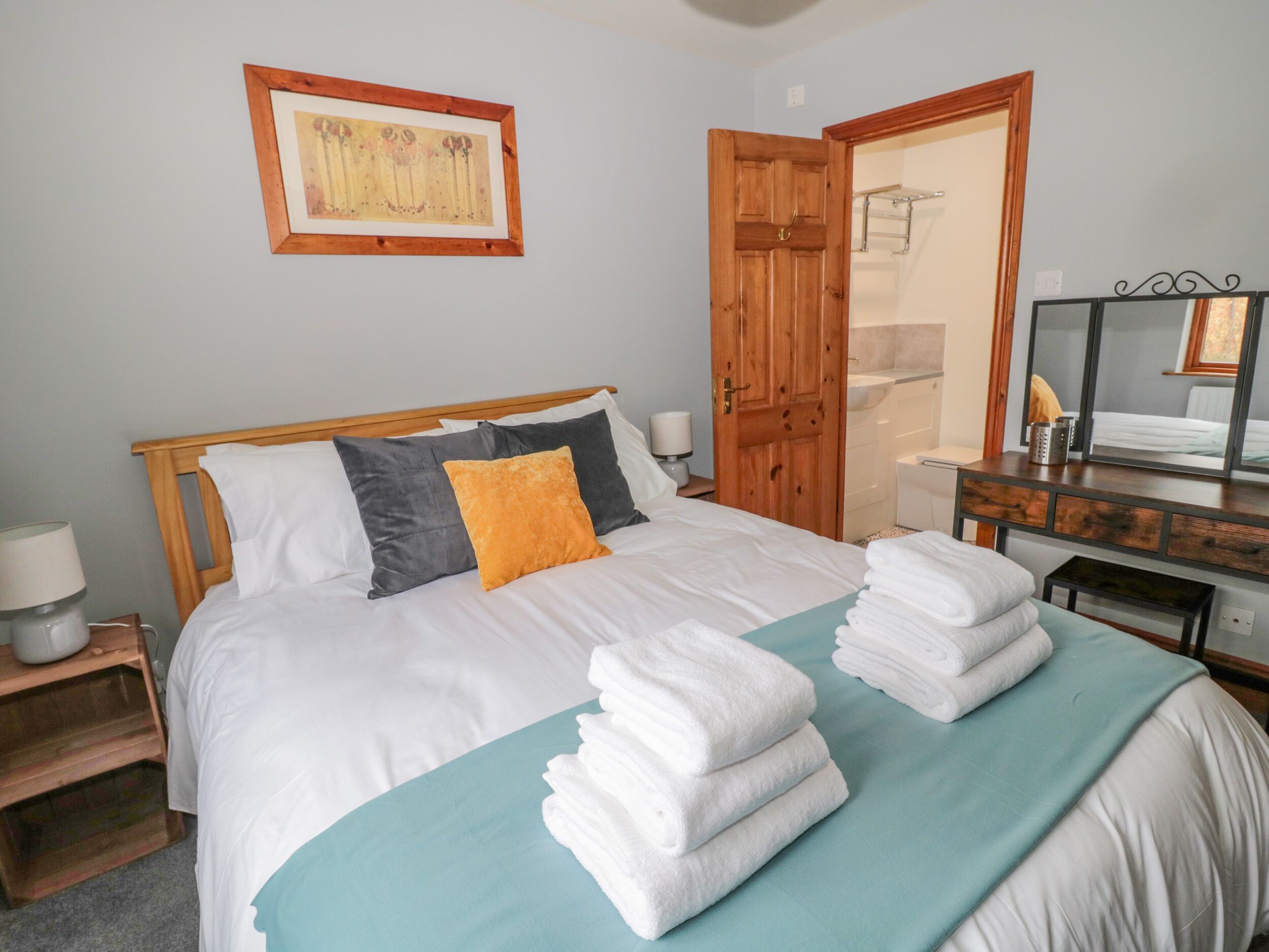 A neatly made bed with white and gray bedding, a teal throw blanket, and two folded white towels. A wooden door and a mirror are visible in the background.