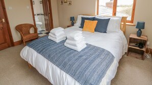 A neatly made bed with a blue and white patterned bedspread, two pillows, and folded towels. There is a wicker chair, a small wooden nightstand with a lamp, and a window with a blue-striped valance. - Ridley Lodge (Morpeth)