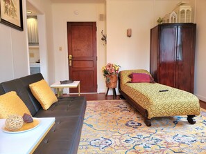 Ein Zimmer mit einem dunkelbraunen Sofa, einem kleinen Tisch mit einer Dekoration, einem Bett mit gemustertem Überwurf, einem Holzschrank und einem Teppich mit Muster.