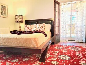 Un lit bien fait, agrémenté d’oreillers à motifs floraux, d’une lampe de chevet et d’un tapis rouge à motifs.