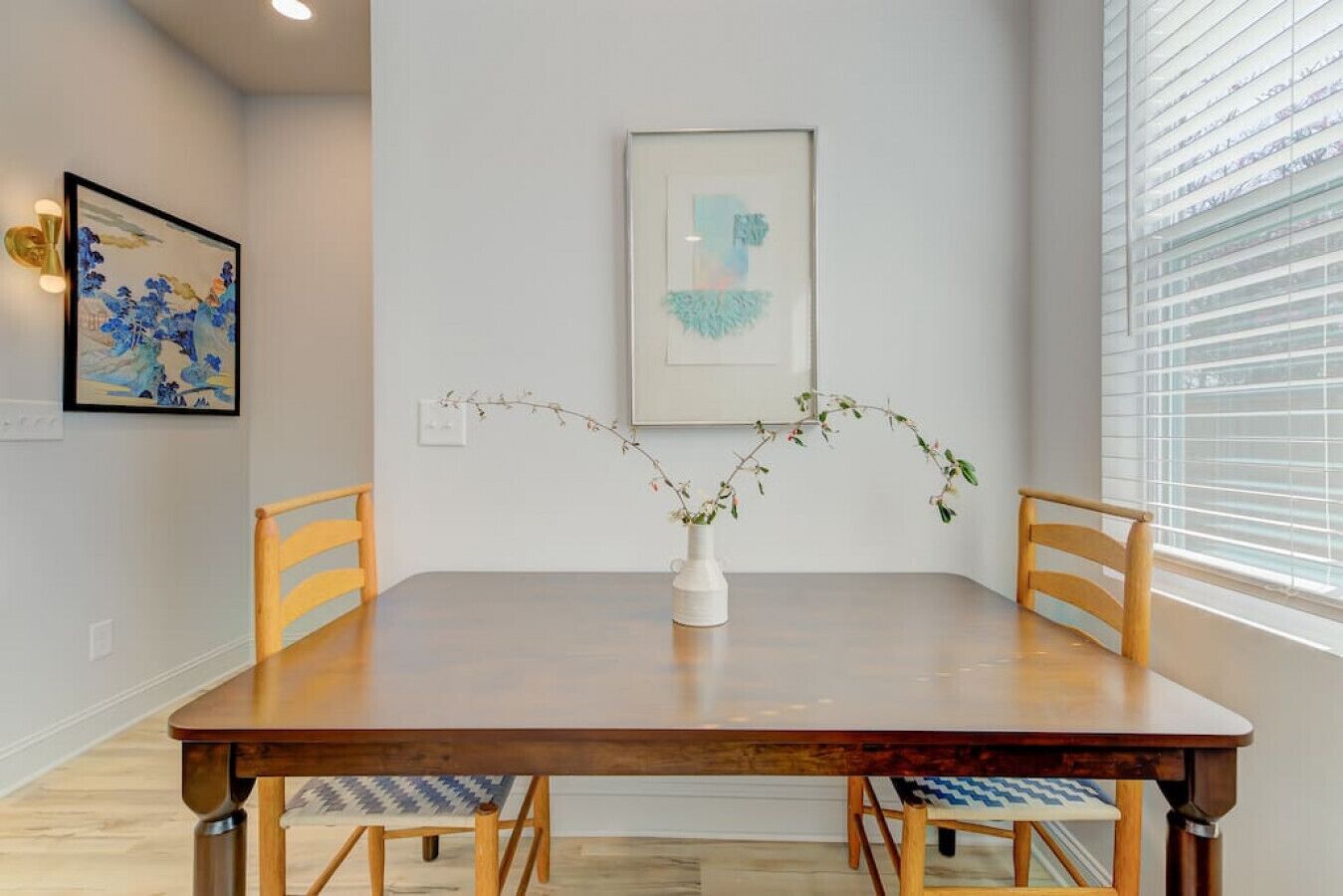 A dining area with a wooden table, two chairs, a vase with a plant, and two framed artworks on the wall.
