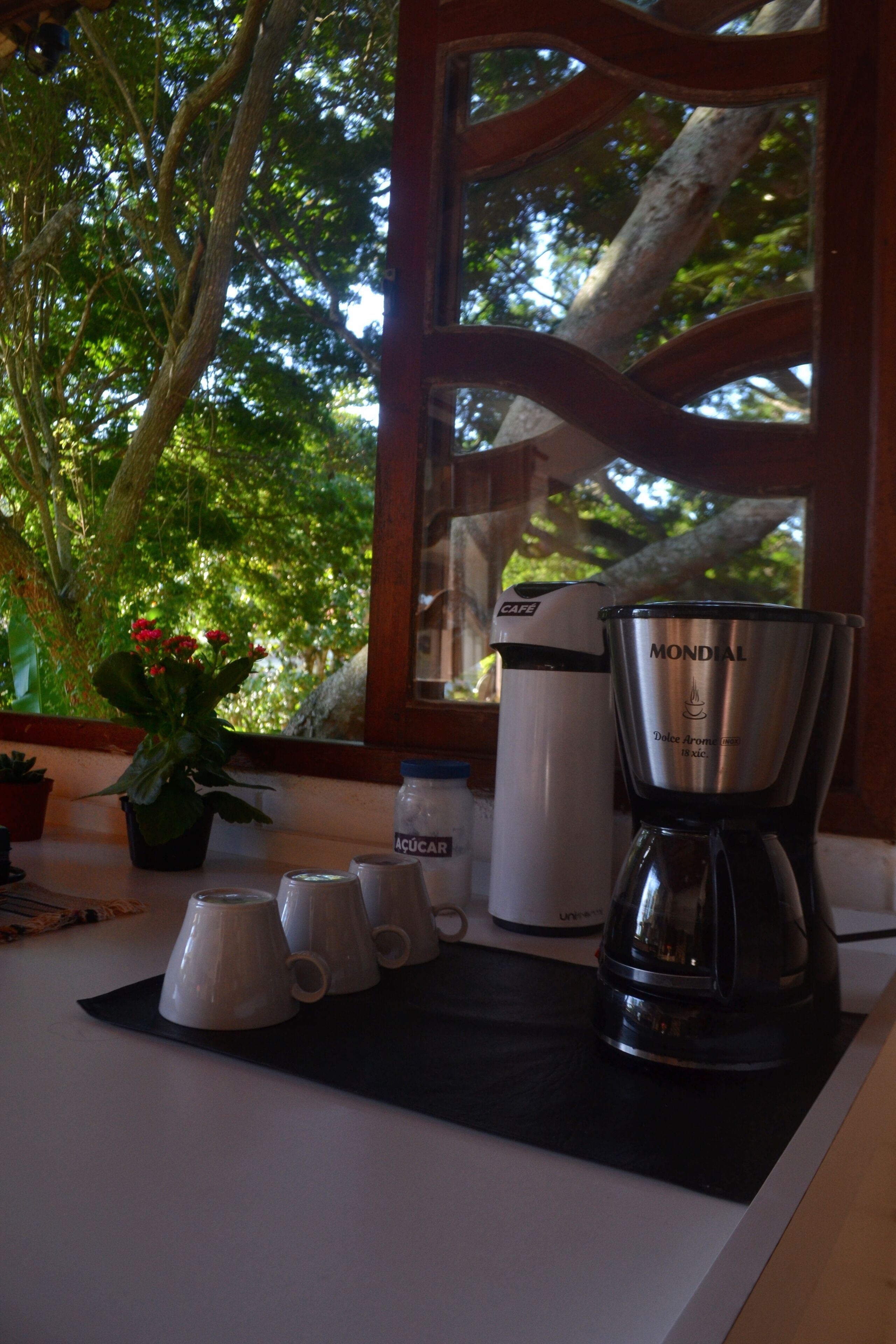 A coffee maker, a sugar container, and four cups on a kitchen counter near a window with a view of trees.