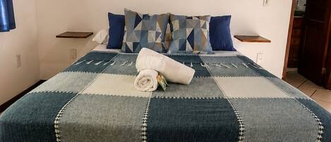 Un dormitorio con una cama grande, techo de madera, repisa con plantas y un cuadro enmarcado.