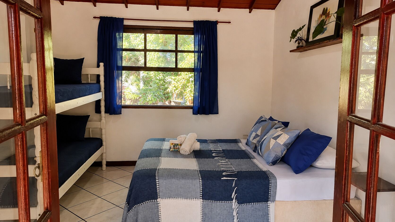 Une chambre avec un lit superposé, un lit simple recouvert d’une couverture à carreaux et une fenêtre avec des rideaux.