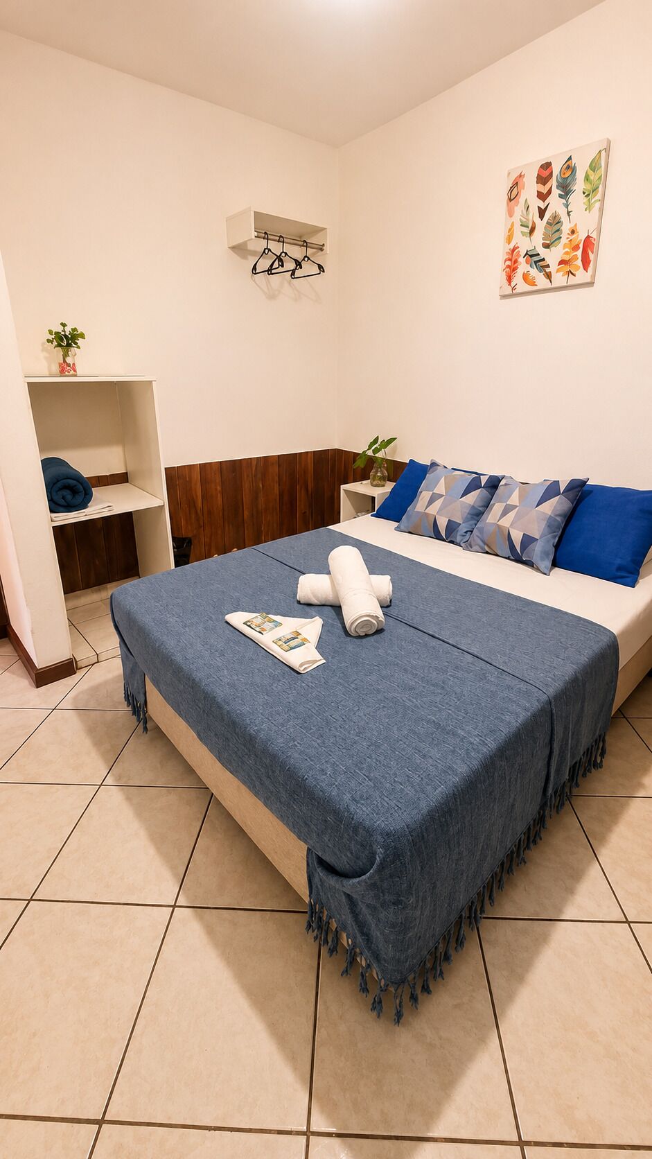 A bedroom with a bed, a bedside table, a wall-mounted air conditioner, a shelf with a plant, and a wall art piece.