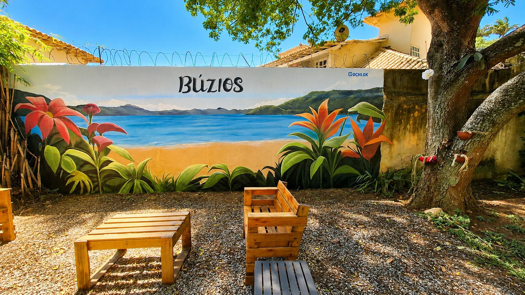 Un mural con una escena de playa, la palabra 'Búzios' y un banco de madera al frente.