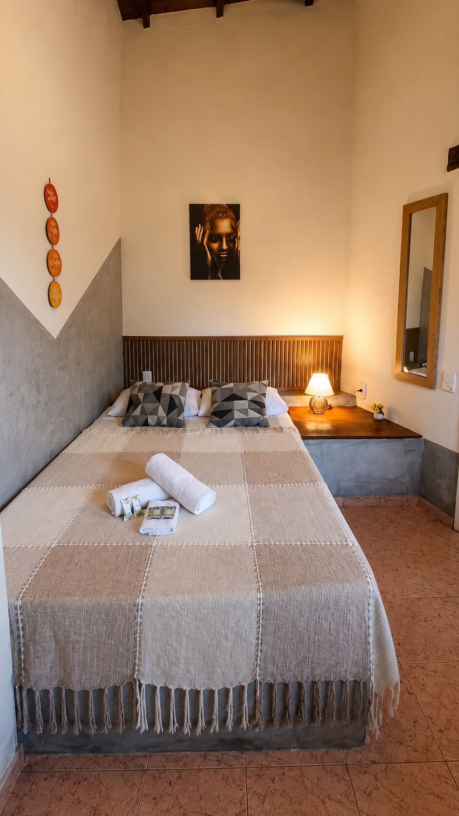 A hotel room with a bed, a bedside lamp, a mirror, and a wall with a decorative item.
