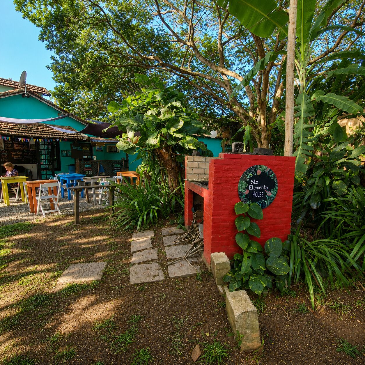 A garden area with a red sign indicating the 5th Element House, surrounded by lush greenery and a building with a tiled roof.