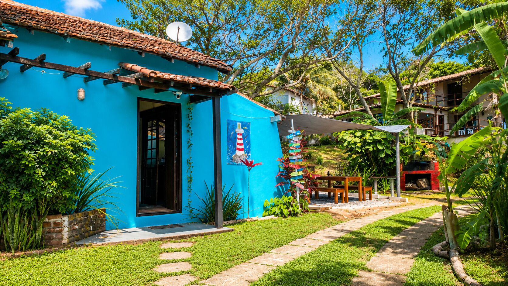 Une maison bleue avec un toit en tuiles, une porte et un petit jardin avec un chemin menant à un espace barbecue rouge.