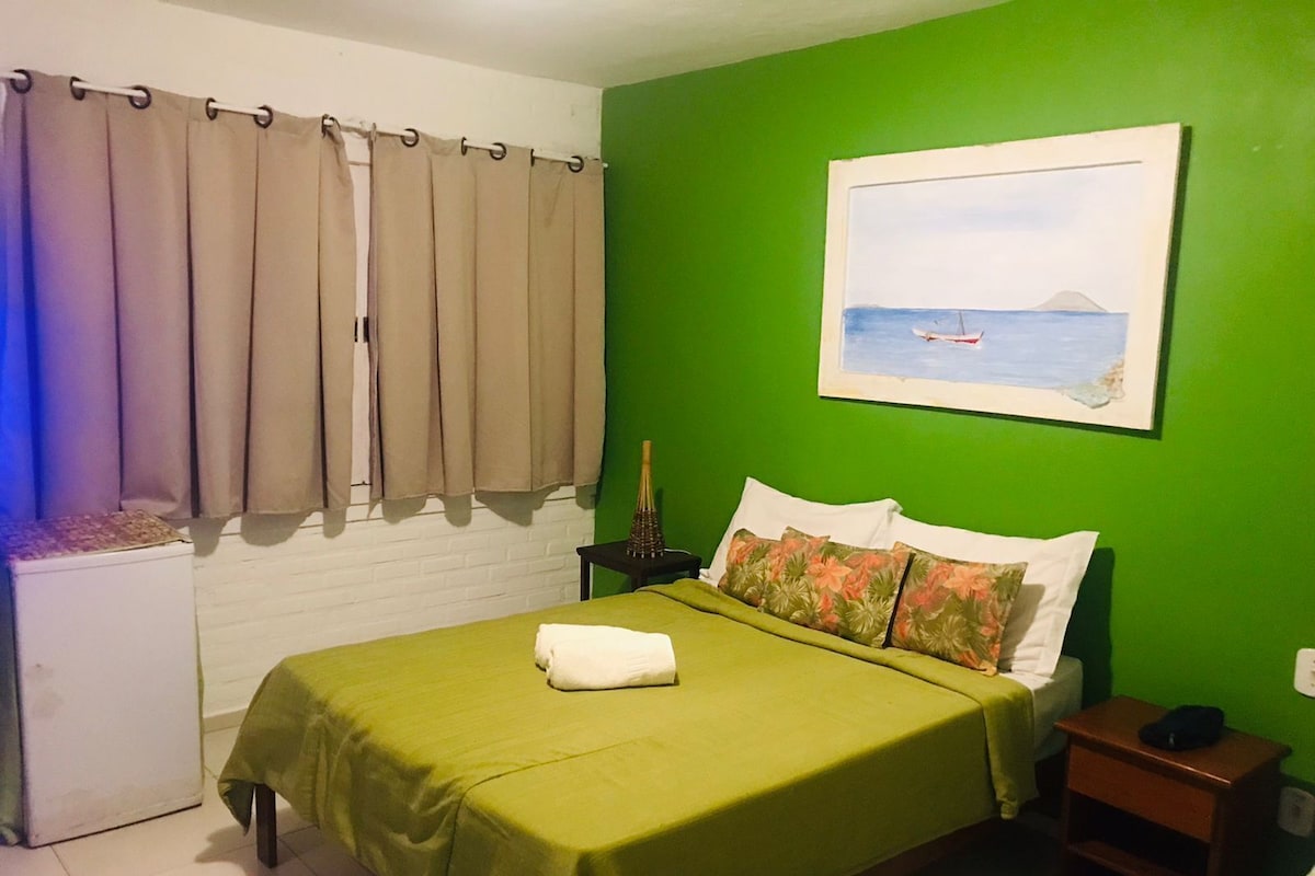 Una habitación con cama, mesita de noche, pared verde, ventana con cortinas y un cuadro.