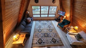 A cozy log cabin interior with a large window, a sofa, a chair, a patterned rug, and mounted animal heads on the wall. - Luther Secluded Riverfront A-Frame (Luther)
