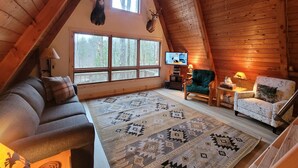 A cozy attic room with a wooden sloped ceiling, a large window, a television, a green armchair, a patterned rug, and a sofa. - Luther Secluded Riverfront A-Frame (Luther)