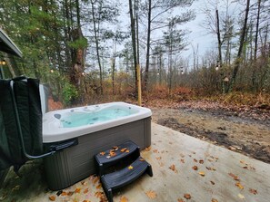 Un bain à remous extérieur doté d’un banc en bois et d’un porte-serviettes, entouré d’arbres et de feuilles mortes.
