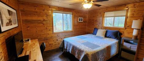 A cozy cabin room with a wooden interior, a bed with patterned bedding, a desk, and a ceiling fan.