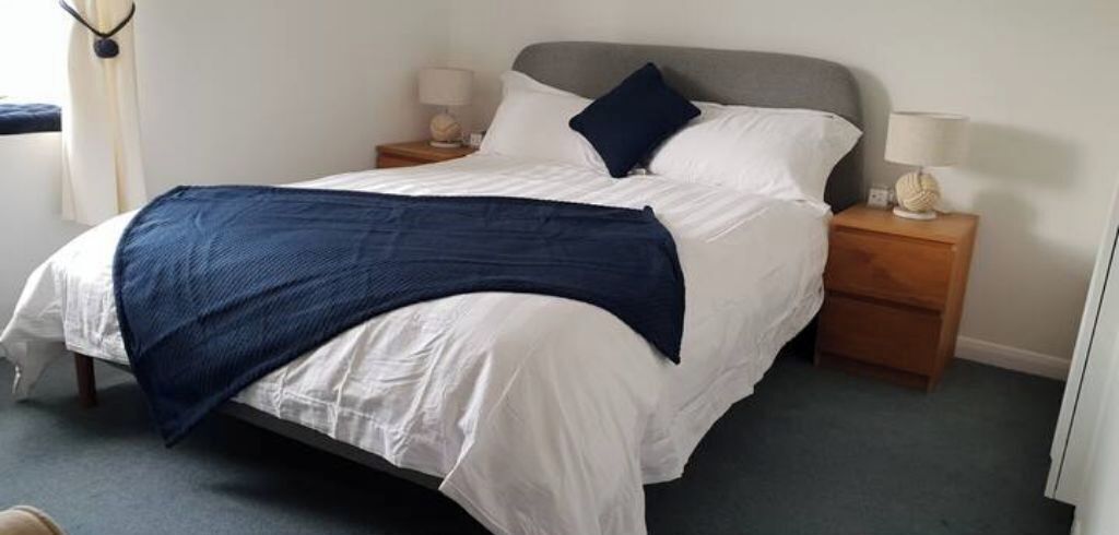 Ein ordentlich bezogenes Bett mit weißer Decke und blauer Tagesdecke. Zwei Nachttische mit Lampen und einem kleinen dekorativen Gegenstand stehen auf jeder Seite. Der Raum hat ein schlichtes, sauberes Design mit grauem Kopfteil und weißen Wänden.